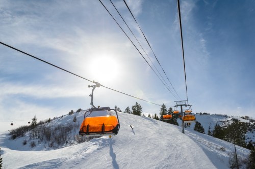 A ski lift with orange gondolas ascends a snowy hill under a bright sun and blue sky.