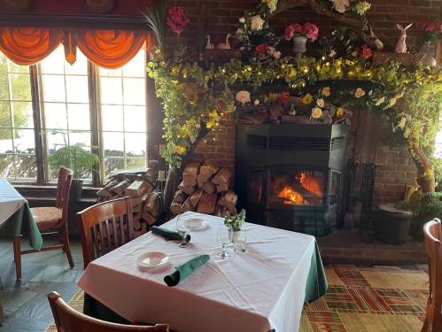 Cozy restaurant interior with a fireplace, floral decorations, and tables set for dining. Warm and inviting atmosphere.