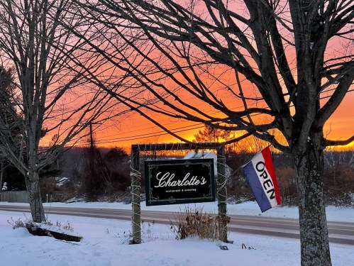 A restaurant sign reading "Charlotte's" with an "OPEN" flag, framed by trees against a vibrant sunset sky.