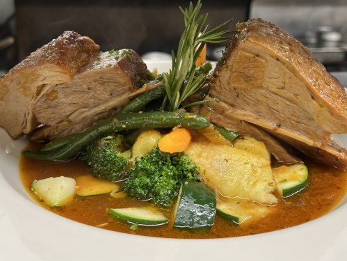 A plate of tender meat with vegetables, including broccoli, zucchini, and green beans, garnished with a sprig of rosemary.