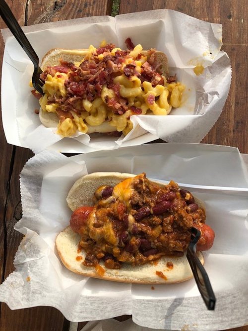 Two hot dogs in paper trays: one topped with macaroni and cheese and bacon, the other with chili and cheese.