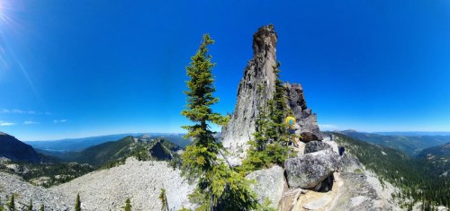 A panoramic view of a rocky mountain peak surrounded by green trees under a clear blue sky.