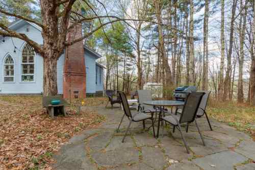 A stone patio with outdoor furniture surrounded by trees, next to a blue house with a unique architectural style.