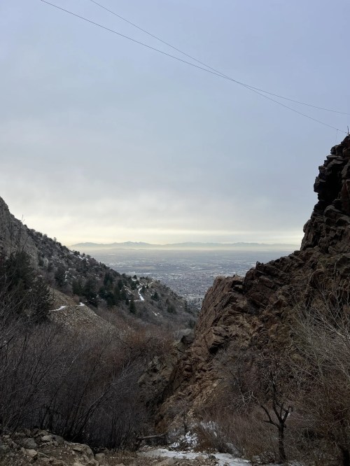 A scenic view of a valley between rocky mountains, with a distant city skyline under a cloudy sky.