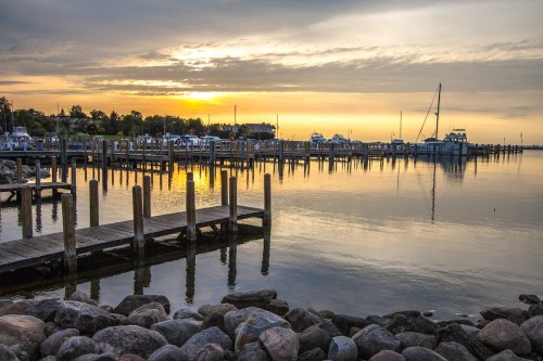 Sunset over a calm harbor with boats docked and a wooden pier extending into the water, surrounded by rocky shore.