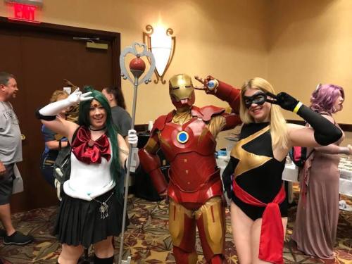 Three cosplayers pose together: one in a sailor outfit, one in an Iron Man suit, and one as a superhero with a mask.