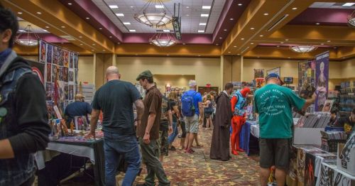 A bustling convention hall filled with vendors and attendees browsing comic books and merchandise.