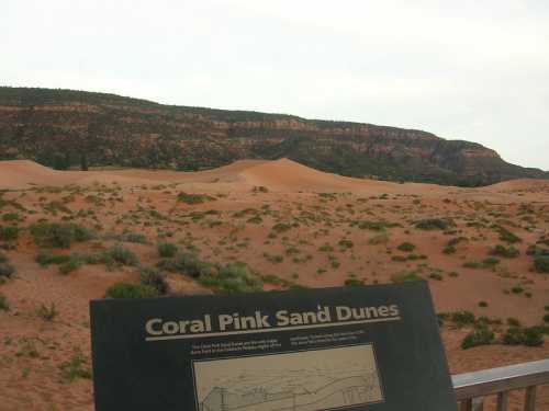 Sign for Coral Pink Sand Dunes in front of sandy dunes and a rocky hillside under a cloudy sky.