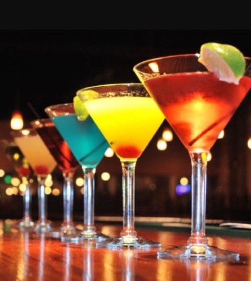 Five colorful cocktails in elegant glasses, lined up on a bar with soft lighting in the background.