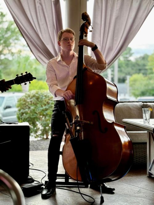 A musician plays a double bass in a cozy setting, wearing a white shirt and black pants, with a guitar nearby.