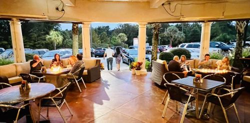 A cozy outdoor seating area with people dining and socializing, surrounded by palm trees and parked cars in the background.