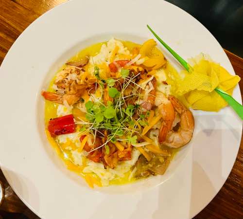 A plate of shrimp and rice topped with vegetables and microgreens, garnished with a yellow flower and green pick.