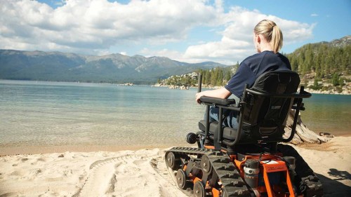 A person in a tracked wheelchair sits on the beach, facing a calm lake and mountains under a blue sky.