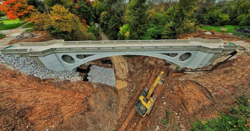 A newly constructed bridge spans a construction site, surrounded by trees and autumn foliage. Heavy machinery is visible below.