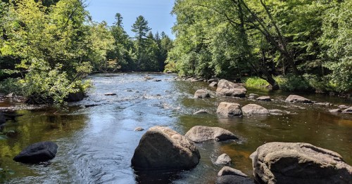 A serene river flows through a lush green landscape, with large rocks scattered along the water's edge.