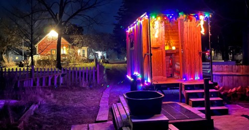 A cozy wooden cabin adorned with colorful lights at night, surrounded by trees and a distant red house.