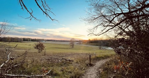 A serene landscape at sunset, featuring a winding path, open fields, and trees silhouetted against the colorful sky.