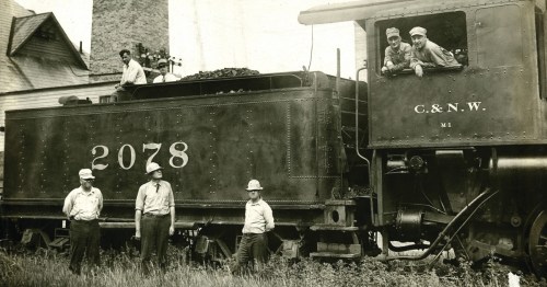 Black and white photo of a vintage train with five men posing, two in the engine and three standing beside it.