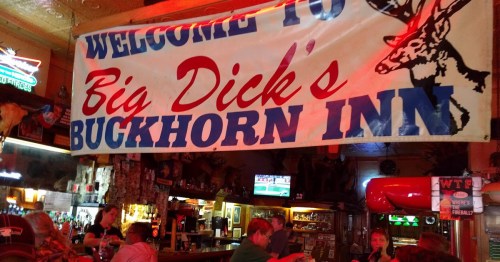 A colorful banner reading "Welcome to Big Dick's Buckhorn Inn" hangs above a lively bar scene with patrons.