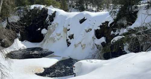 A snowy landscape featuring a frozen waterfall cascading into a dark pool, surrounded by trees and rocky cliffs.