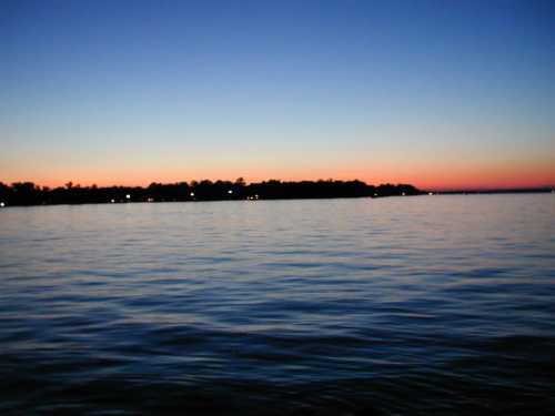 A serene twilight scene over calm water, with a colorful gradient sky and silhouettes of trees along the horizon.