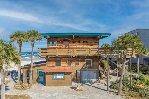 A wooden beach house with a balcony, surrounded by palm trees, near the ocean under a blue sky.