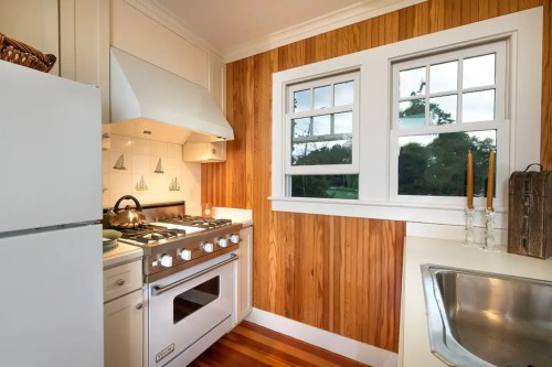Cozy kitchen with wooden walls, white appliances, a gas stove, and two windows letting in natural light.
