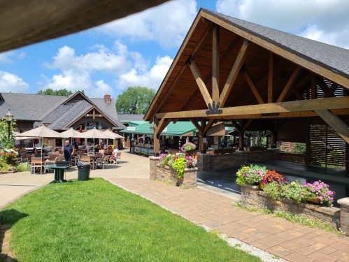 A sunny outdoor dining area with wooden structures, tables, and colorful flowers, surrounded by green grass.