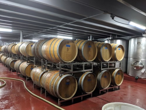 A wine cellar with stacked wooden barrels and stainless steel tanks, illuminated by overhead lights.