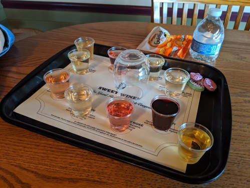 A tray with various wine samples in small cups, a glass, and a water bottle, on a wooden table.