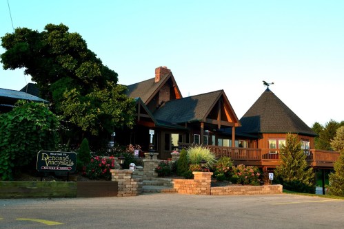 A charming winery building with a wooden exterior, surrounded by greenery and flowers, under a clear blue sky.