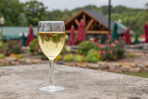A glass of white wine sits on a stone table, with a blurred outdoor setting and colorful umbrellas in the background.