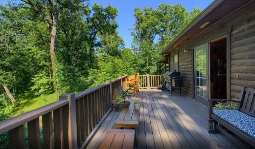 A wooden deck with seating and a grill, surrounded by lush green trees under a clear blue sky.