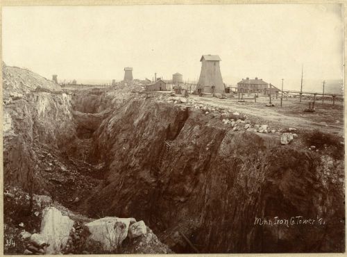 Historic black-and-white photo of a mining site with structures and a large excavation area.