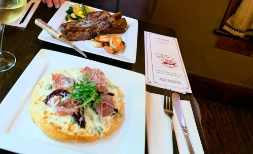 A table setting with a steak, shrimp, vegetables, and a creamy dish topped with prosciutto and greens.