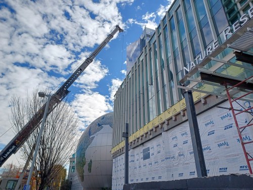 A construction crane works on a modern building with a globe sculpture outside, under a partly cloudy sky.