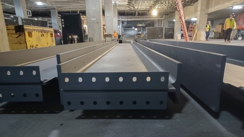 Close-up of steel beams on a construction site, with workers and machinery in the background.