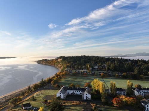 Aerial view of a serene landscape with a river, autumn trees, and historic buildings under a clear blue sky.