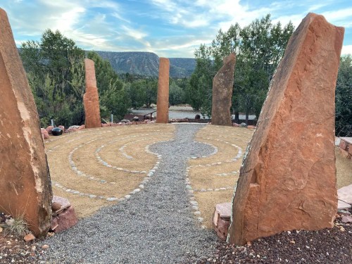 A stone labyrinth surrounded by tall rock formations and trees, set against a scenic landscape and blue sky.