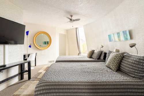 A modern hotel room with two beds, a TV, a round mirror, and large windows letting in natural light.