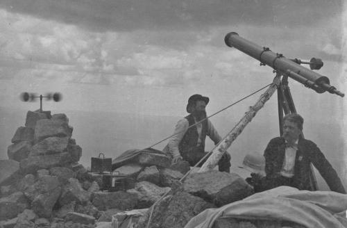 Two men sit on a rocky mountaintop, one operating a large telescope, with cloudy skies in the background.