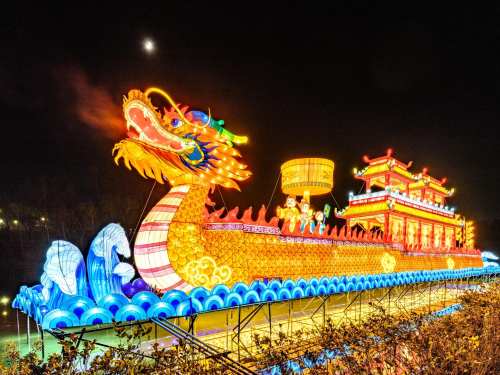 A brightly lit dragon boat sculpture with colorful decorations, set against a night sky with a visible moon.