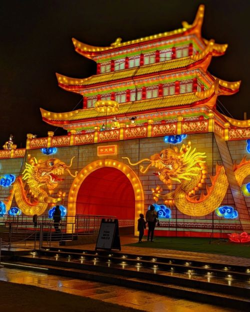 A brightly lit traditional Chinese archway with dragon decorations, set against a night sky. People stand nearby.