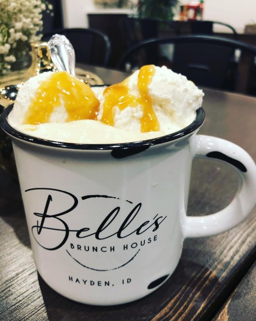 A white mug with "Belle's Brunch House" logo filled with creamy dessert topped with whipped cream and caramel sauce.