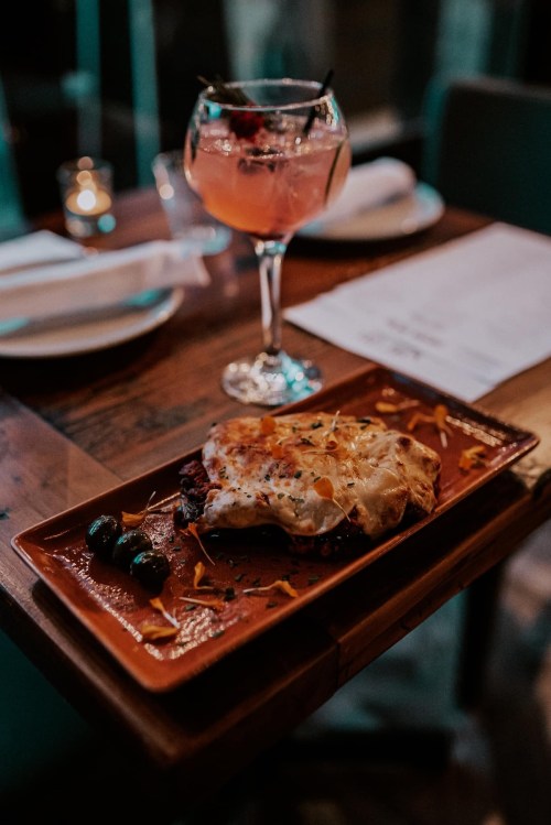 A plate of lasagna topped with cheese and herbs, accompanied by a cocktail in a glass, on a wooden table.