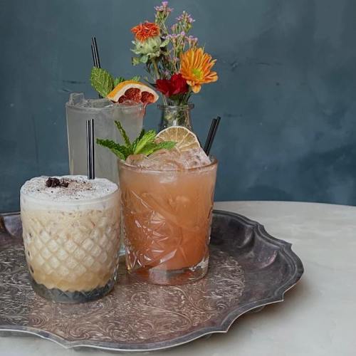 Three colorful cocktails on a silver tray, with a small vase of flowers in the background.