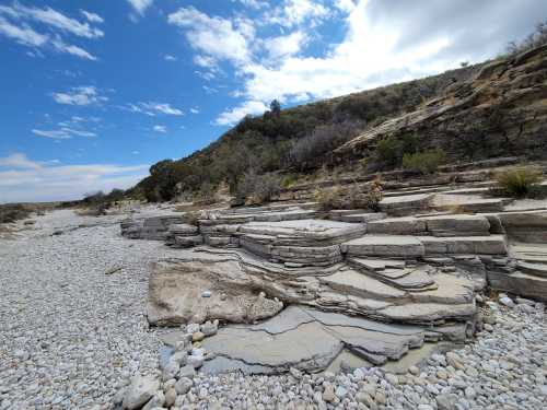 Rocky landscape with layered stone formations and a gravelly riverbed under a partly cloudy sky.