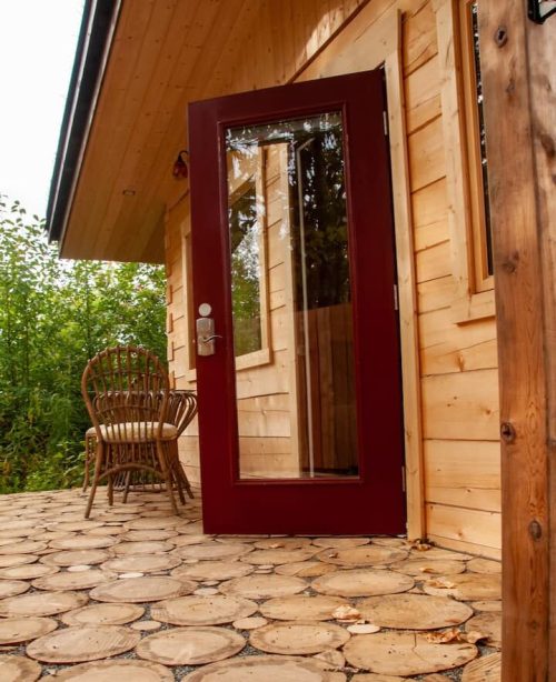 A wooden cabin entrance with a red door, surrounded by greenery and a stone pathway leading to a chair.