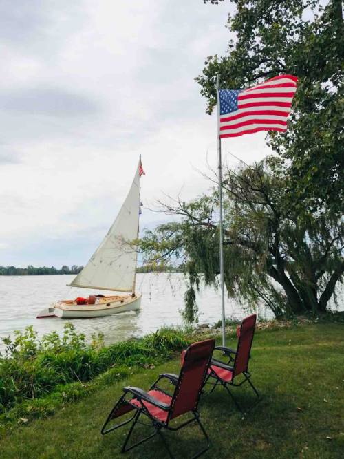 A sailboat on a lake near a flagpole with an American flag, with two empty chairs on the grassy shore.