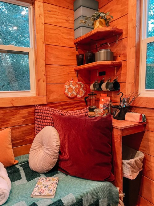 Cozy corner of a wooden cabin with cushions, a small table, and kitchenware on shelves, surrounded by natural light.